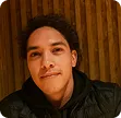 Portrait of a young man with curly hair wearing a dark jacket against a wooden background.